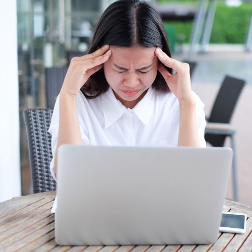 Mujer asiática con dolor de cabeza sentada en una mesa al aire libre con una computadora portátil buscando dentista general.