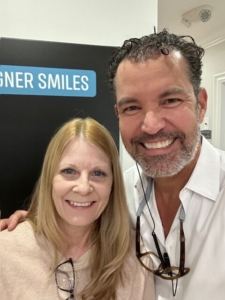 Two smiling individuals posing for a selfie in front of a sign that reads "designer smiles".