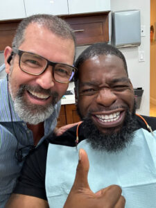 Two men smiling at the camera, one with glasses, in a dental office setting, with the patient wearing a dental bib and giving a thumbs up.