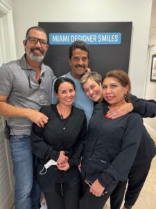 A group of five people, three women and two men, stand smiling in front of a sign that reads "Miami Designer Smiles". They are wearing casual and professional attire.