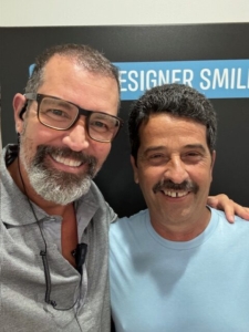 Two men are standing close to each other and smiling. The man on the left has glasses and a beard, while the man on the right has a mustache. They are in front of a black wall with partially visible text.