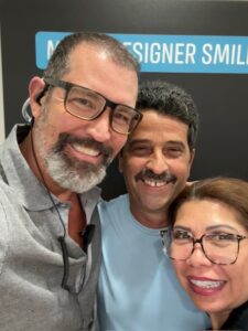 Three people are smiling for a selfie in front of a sign that partially reads "DESIGNER SMIL." They are wearing glasses and casual clothing.