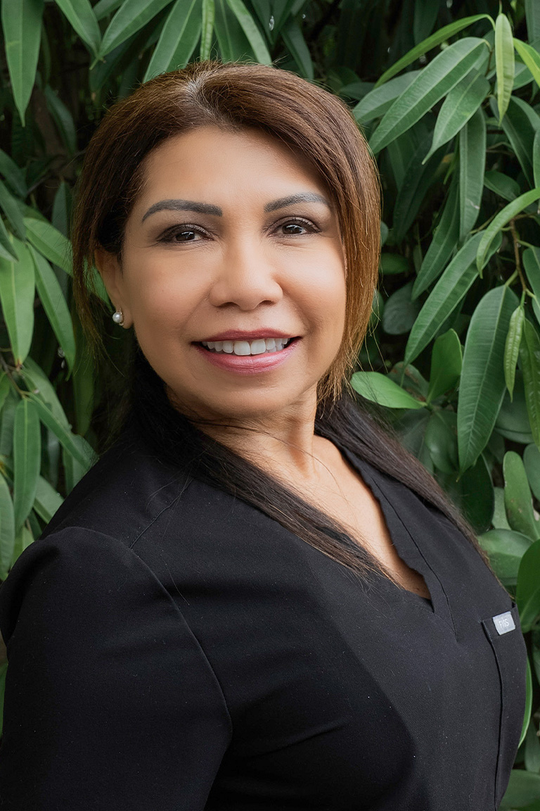 Woman with long dark hair wearing a black top, smiling while standing in front of green leafy plants, embodies the welcoming spirit often found in a dedicated dental team.