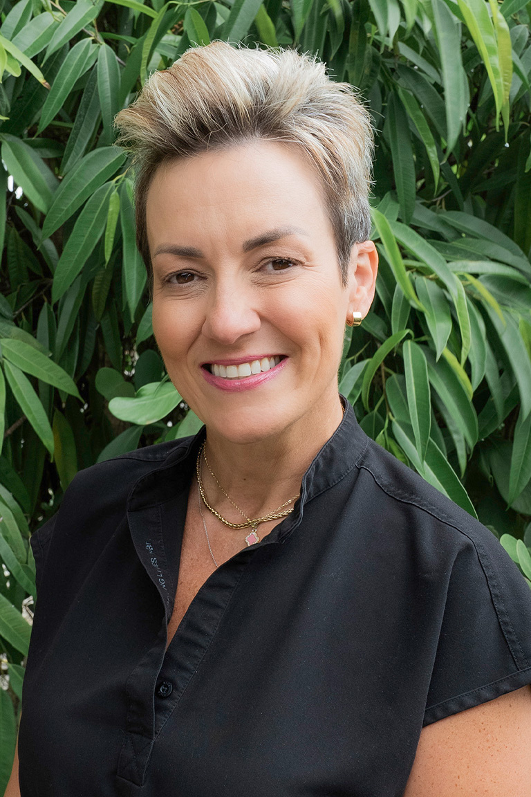 A woman with short, light brown hair wearing a black collared shirt stands smiling in front of green leafy plants, representing the Dental Team.