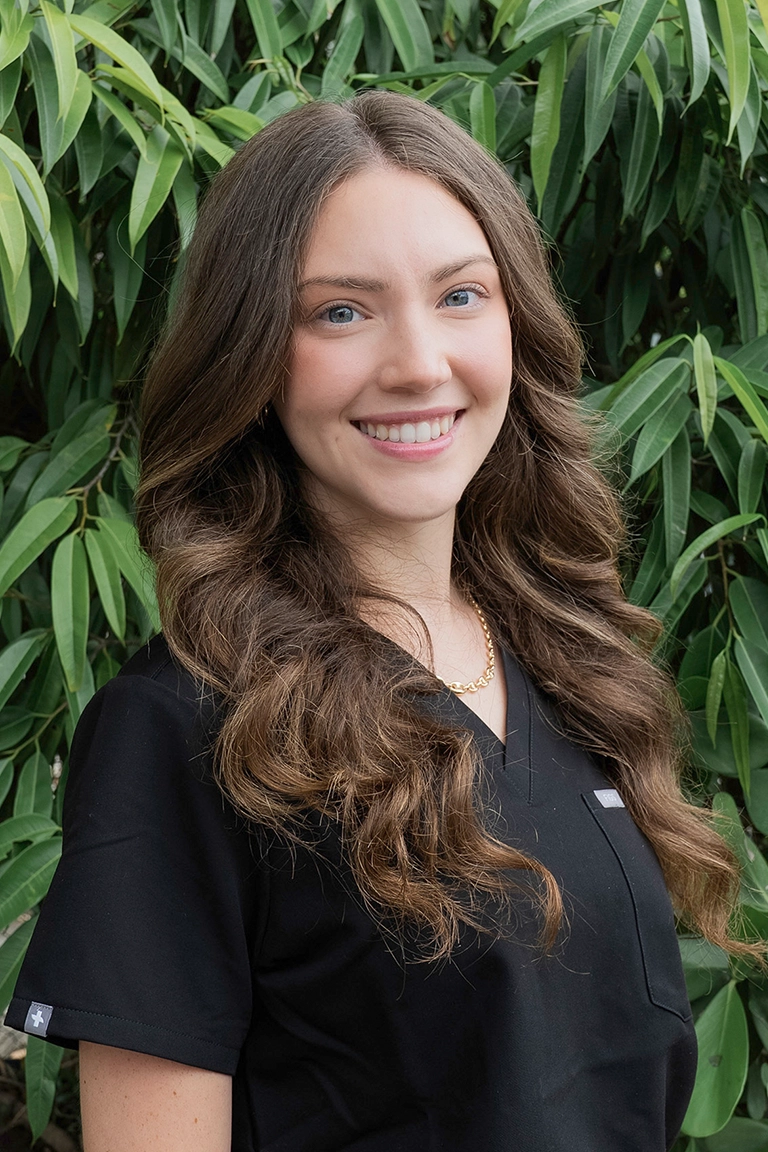 A woman with long wavy brown hair, wearing a black top and gold necklace, stands smiling in front of green foliage—her warm demeanor reflecting the approachable spirit of dedicated dental professionals.