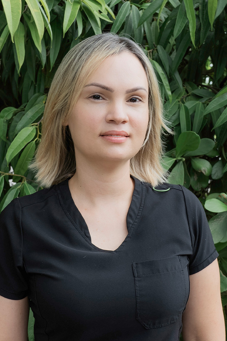 A woman with short blonde hair wearing a black shirt stands in front of green leafy foliage, looking at the camera with a neutral expression—an approachable member of our dental team.