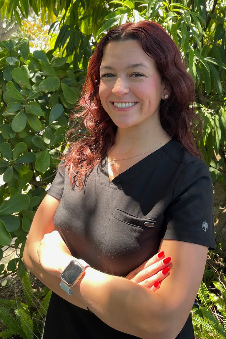 A woman with wavy brown hair, wearing a black scrub top and smartwatch, stands outdoors with arms crossed and smiles confidently, representing the Dental Team in front of green foliage.