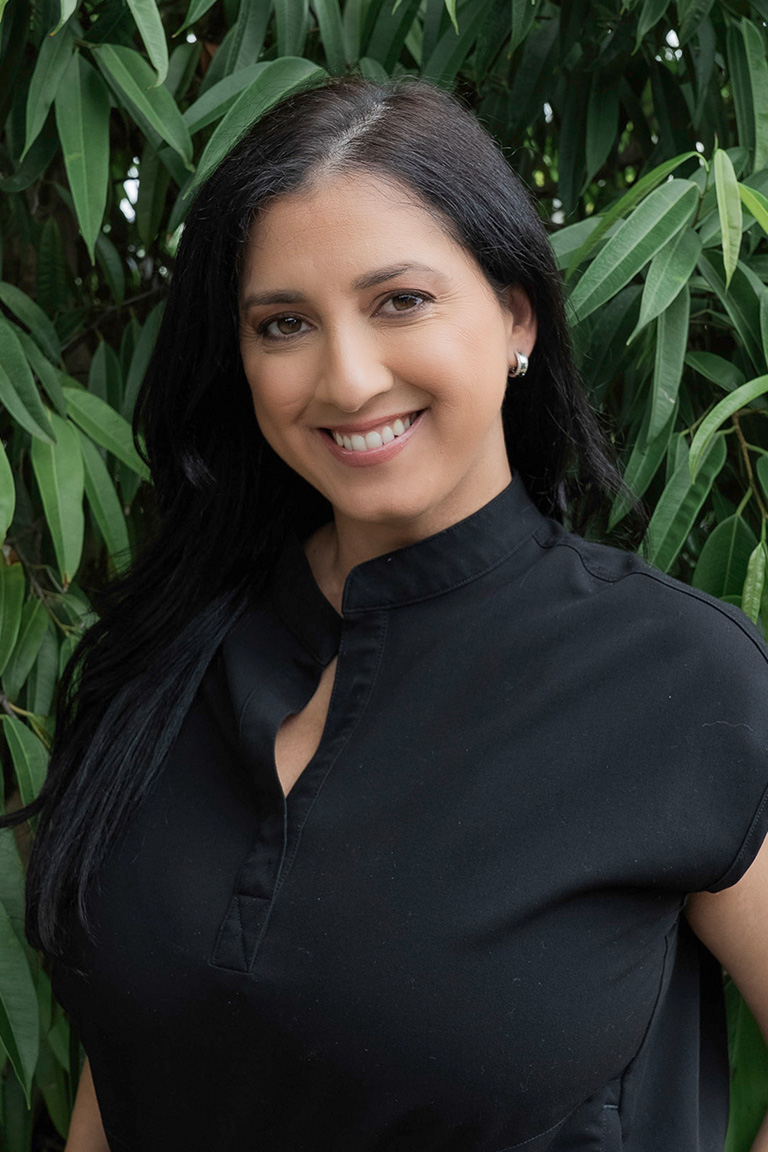 A woman with long black hair wearing a black top stands in front of green foliage, smiling at the camera, radiating the warmth and confidence often seen among dedicated dental professionals.
