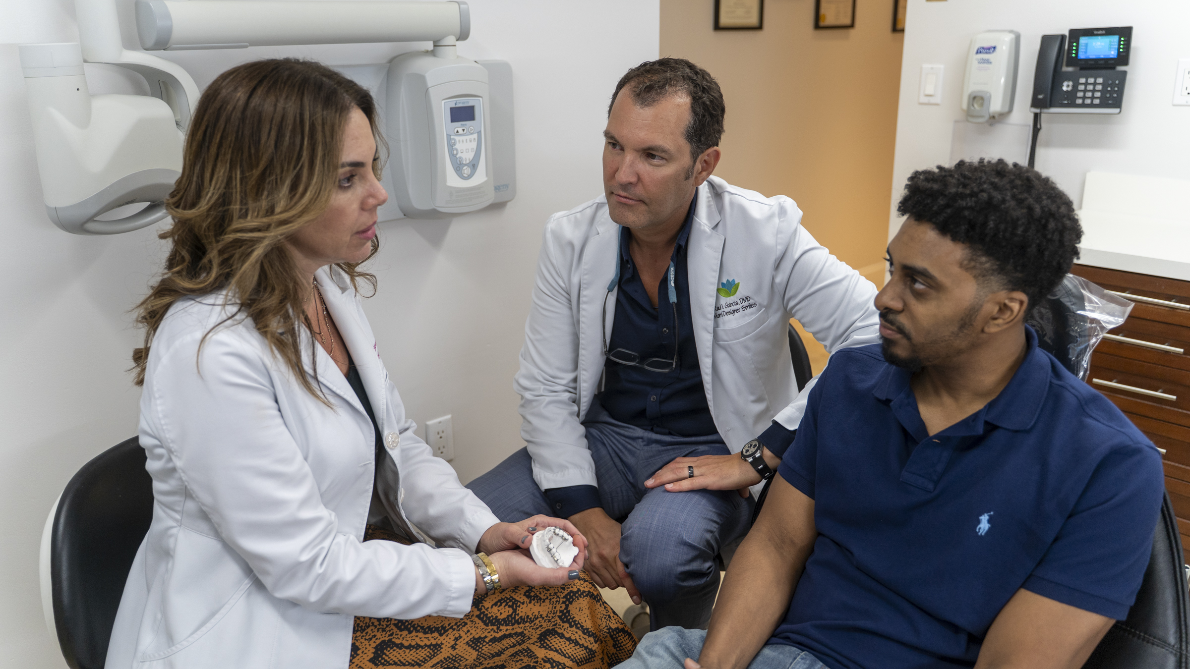 Two doctors in white coats discuss MARPE and traditional orthodontics treatment options with a male patient in a clinic exam room.