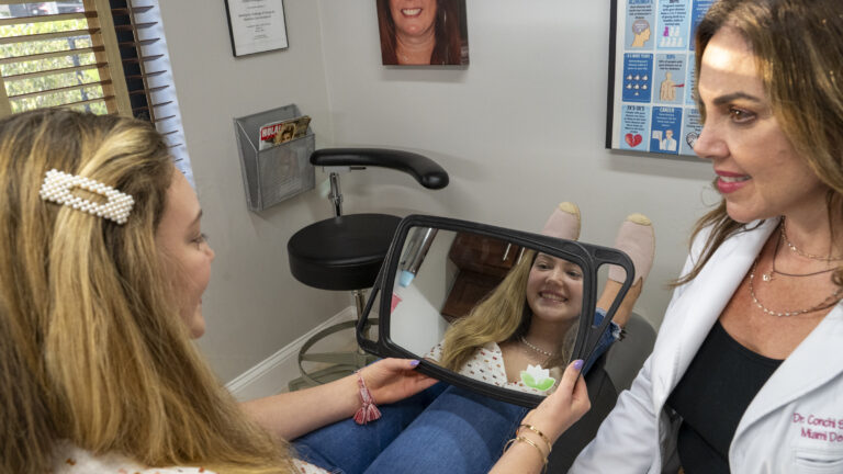 A woman sits in a Miami Cosmetic Dentistry office holding a mirror and smiling at her reflection while a female doctor in a white coat sits nearby.