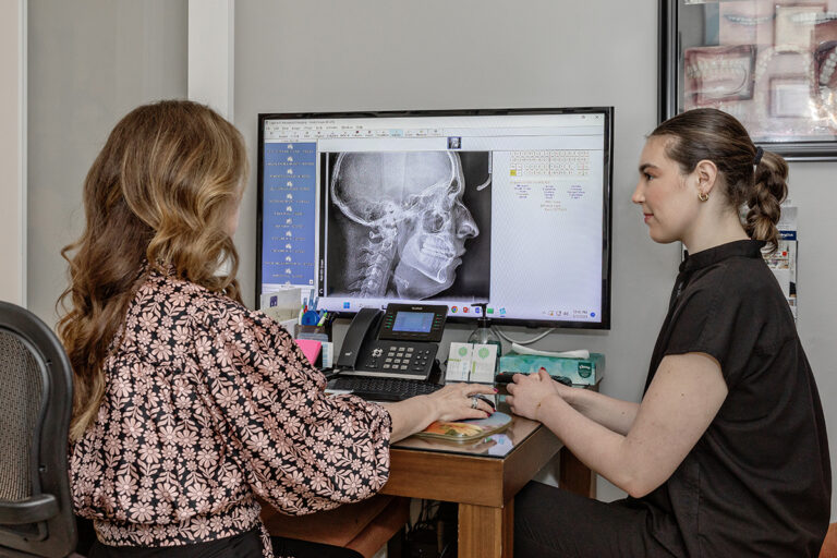 Two women sit at a desk in a Miami Cosmetic Dentistry office, reviewing a side-view X-ray of a skull displayed on a computer monitor, possibly discussing MARPE cost options.