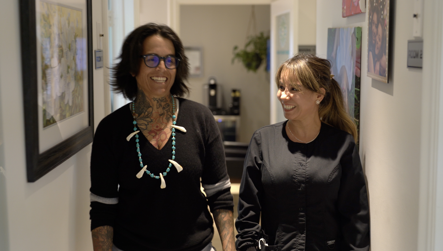 Dos mujeres caminan por un pasillo, sonriendo y conversando. Una lleva tatuajes visibles y gafas, mientras que la otra viste un uniforme negro. De las paredes que las rodean cuelgan obras de arte.