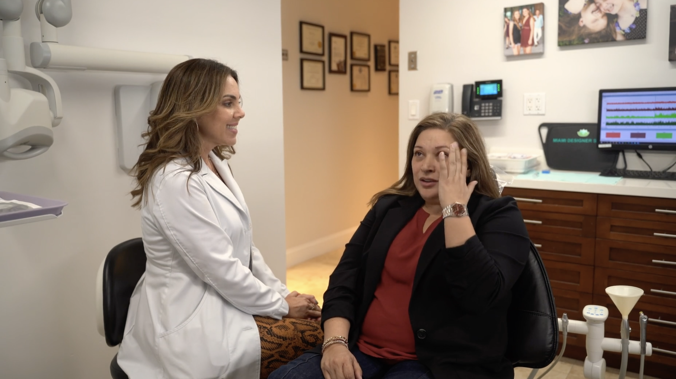 A woman in a black blazer gestures to her face while seated in a medical office, discussing migraine relief with a healthcare professional in a white coat.