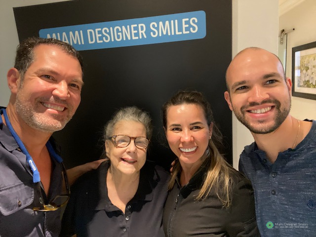 Four people smiling and posing together in front of a sign that reads “Miami Designer Smiles” inside a well-lit room, celebrating the arrival of a new patient.